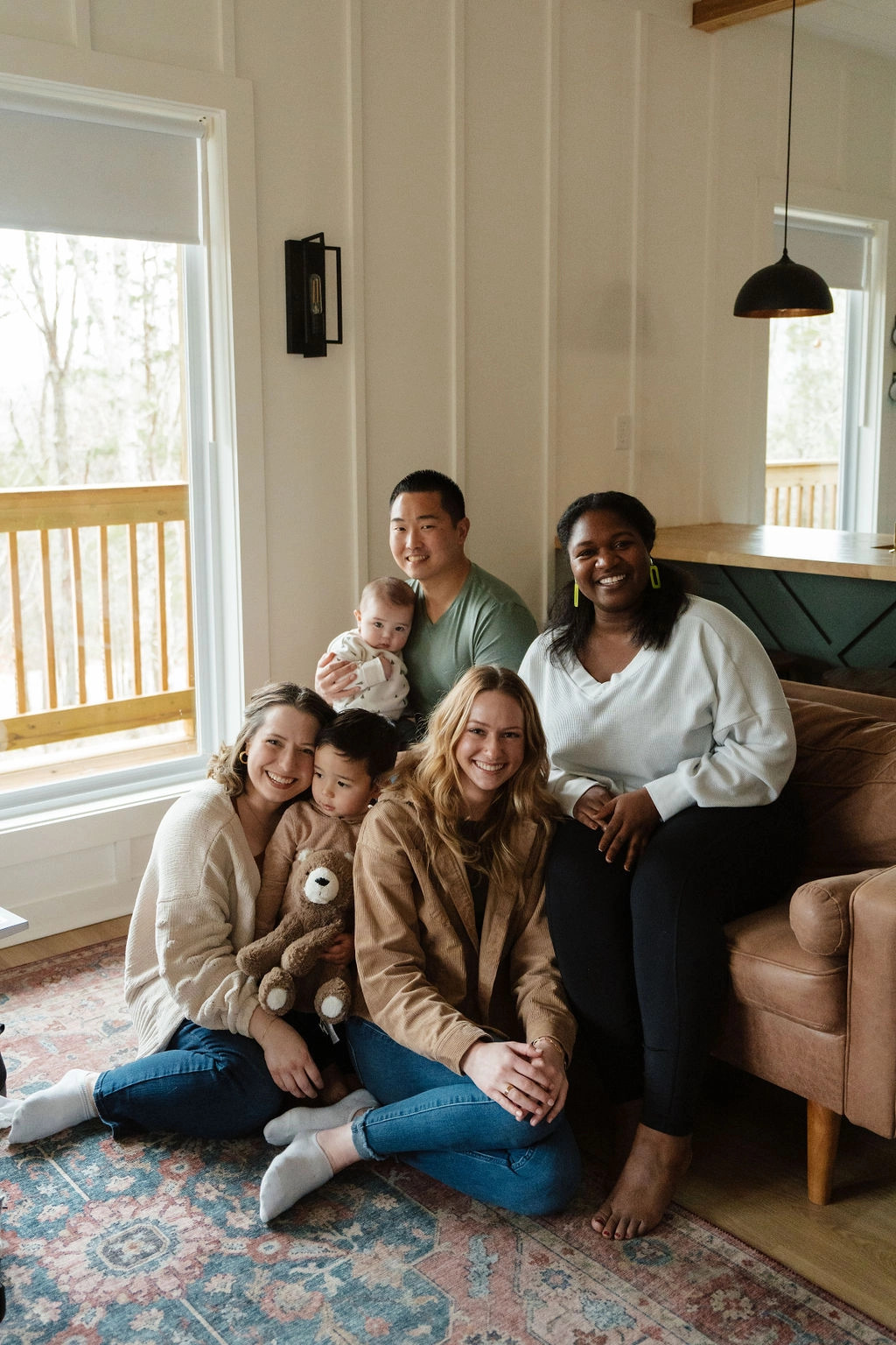 a photo of Amanda Chong, from Appmanda, and her family. The family is sitting on and around a brown leather couch. From right to left: Amanda, on the ground, with her son, William in her lap (around 3 years old), Cassie, on the ground, Jessy, sitting on the couch, behind them sit, Eddie, Amanda's husband. Eddie is holding the baby, David. Amanda is wearing a cream sweater, blue jeans, socks, and various jewelry. William is wearing an oat colored sweater, dark pants, and holding a brown-colored bear. Cassie is wearing a medium brown jacket over a dark tank top with blue jeans, socks, and various jewelry. Jessy is wearing an off-white sweater, black pants, and neon green earrings. Eddie is wearing a green top. His bottom half is hidden. David is wearing a white sweater with small green trees on it. Below them is a rug. Behind them are several windows through which you can see the railings of a porch. Off to the right you can also see a green kitchen cabinet with a wood countertop with a black pendant hanging above the counter.