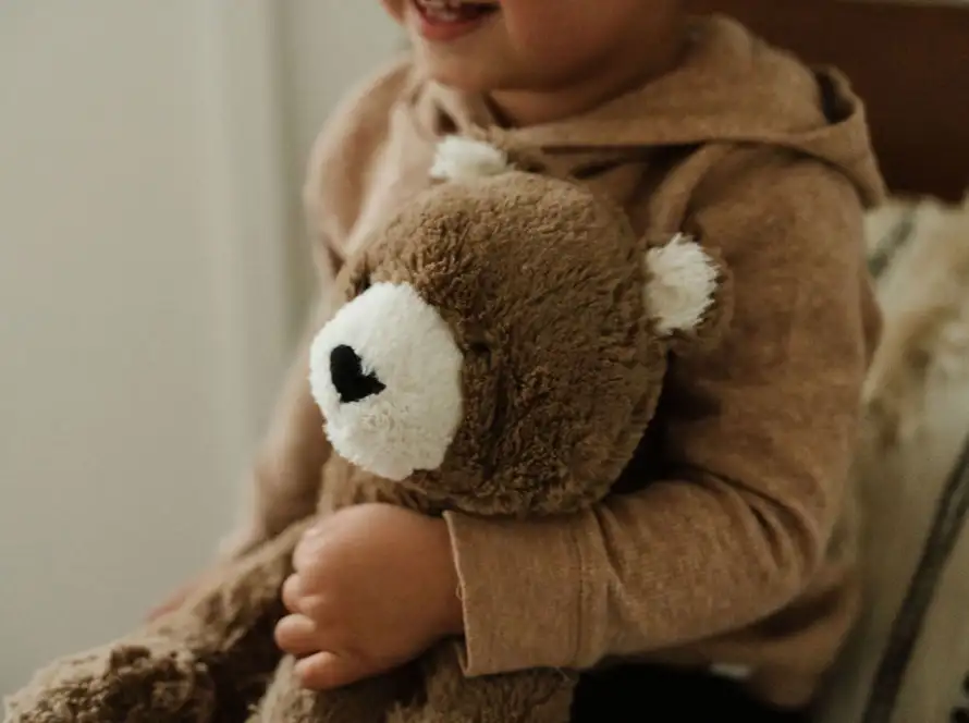 A closeup photo of William Chong holding his favorite stuffed animal, Bear. The teddy bear is a brown bear with white muzzle and ear. William is wearing an oat-colored top and sitting on a chair.
