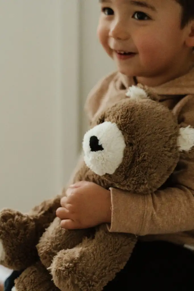 A closeup photo of William Chong holding his favorite stuffed animal, Bear. The teddy bear is a brown bear with white muzzle and ear. William is wearing an oat-colored top and sitting on a chair.