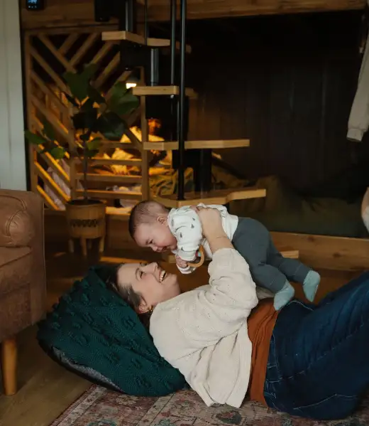 a photo of Amanda Chong, from Appmanda, playing with her youngest son. She is laying on the ground with a dark green pouf behind her head and she is holding her baby up over her. Both Amanda and the baby are smiling. Amanda is wearing an orange tank top, dark pants, and a cream sweater. She is also wearing a necklace, bracelet, and earrings. Her son is wearing a white sweater with small green trees on it and blueish-green pants and socks. On the floor is a decorative rug and next to them is a brown leather couch. Behind them is a plant, a spiral staircase, and bunk beds. Out of focus in the background, some people sit on the bed.