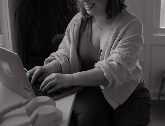 a cropped black and white photo of Amanda Chong sitting at a table and typing on a macbook. Next to her on the table sit a pair of Sony over-the-ear headphones. Amanda is wearing a medium-toned tank top, cream sweater, and dark pants. Behind her is a small stool to the right and a plant to the left along with two windows in the background.