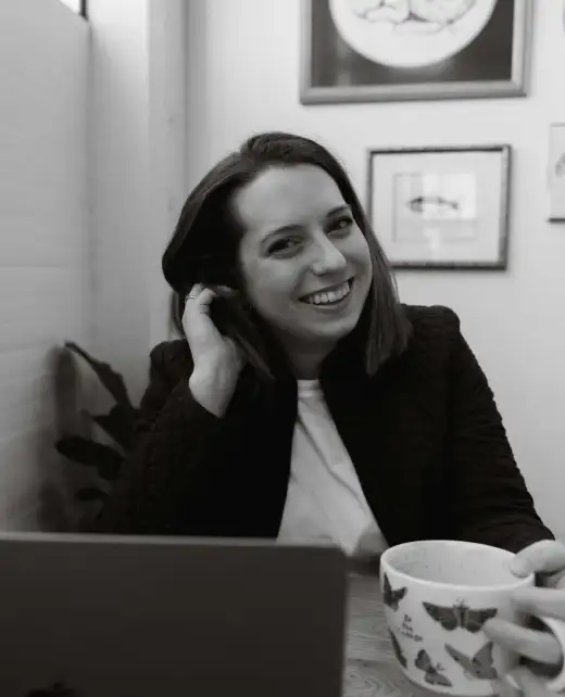 a black and white photo of Amanda Chong sitting at a table and looking at the camera and touching hair with her right hand and holding a butterfly coffee mug in her left hand. In front of her is a gray macbook and a small notebook and pen with notes visible. Amanda is wearing a white shirt and dark jacket. Behind her is a plant and an array of framed images.