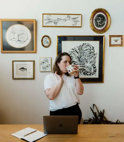 a photo of Amanda Chong, from Appmanda, standing and looking at off camera to the right and touching hair with her right hand and drinking from a butterfly mug in her left hand. Amanda is wearing a white shirt and black pants and a watch on her left wrist. In front of her is a table with a gray macbook and notebook and pen. Behind her is an array of framed artwork and a plant.